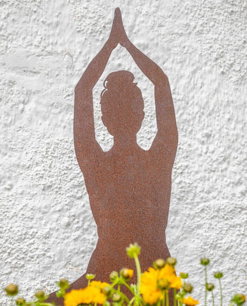 Rostige Metall-Silhouette einer Yoga-Figur zwischen gelben Blumen vor der Hausfassade