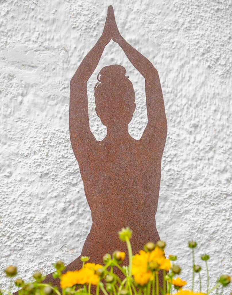 Rostige Metall-Silhouette einer Yoga-Figur zwischen gelben Blumen vor der Hausfassade